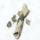 A beige linen napkin with gold stripes and a metallic leaf-shaped napkin ring, arranged on a white marble surface alongside three matching leaf-shaped metal accents.