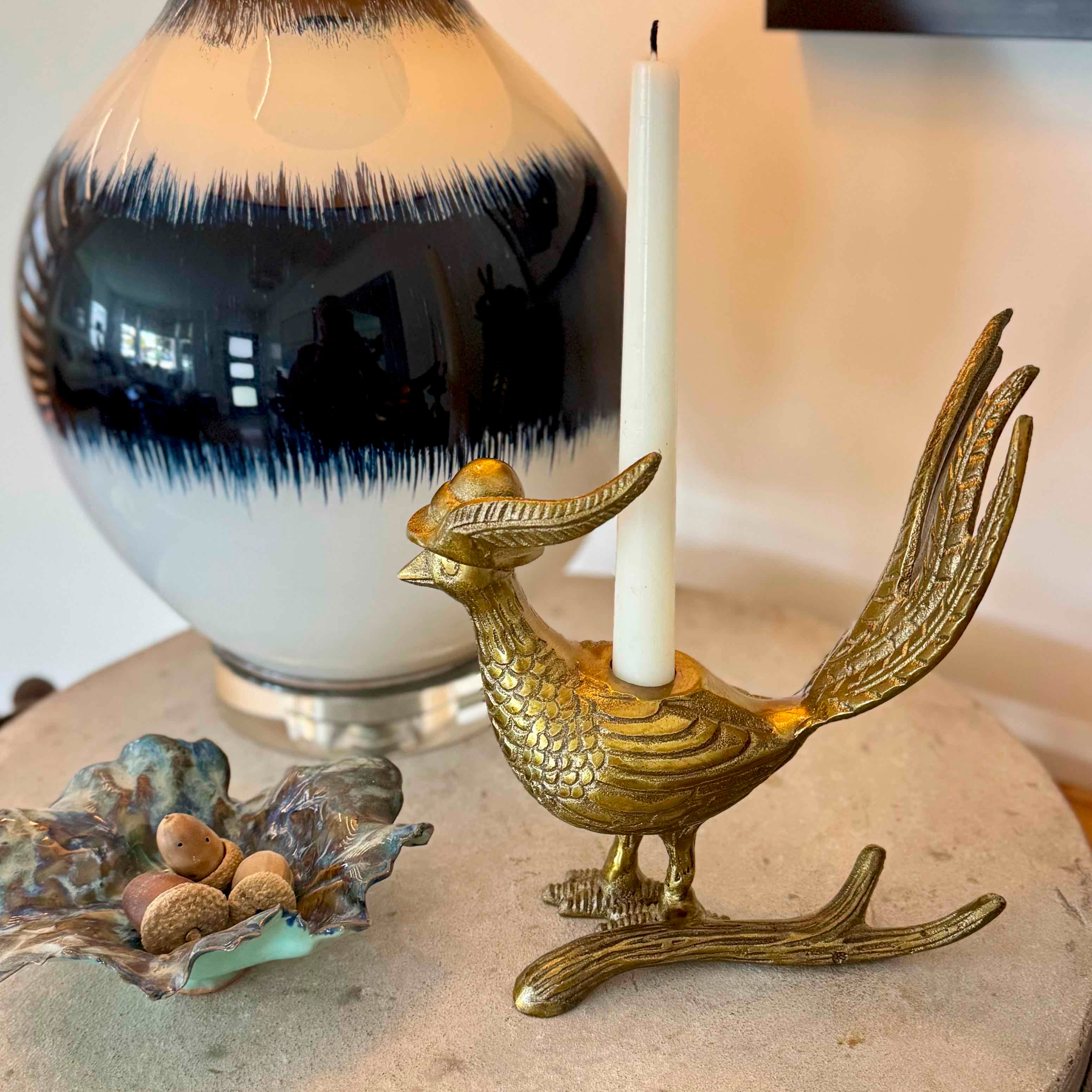 Brass bird-shaped candle holder with a white candle, black and white vase, and decorative shell on a stone surface.