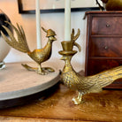 Brass peacock candle holder on a wooden surface with a wooden dresser in the background.