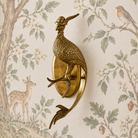 A brass wall-mounted bird fixture shaped like a peacock or pheasant standing on a round backplate, shown against wallpaper with a deer, tree branches, and a small bird.