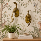 Two ornate gold-toned metal rooster wall mounts with oval backplates, displayed on a cream floral wallpapered wall above a wooden console table with a potted fern and books.