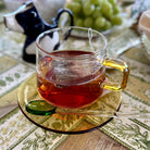 Clear glass mug with yellow handle filled with red tea, placed on a patterned tablecloth.
