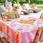 A pink and orange patterned tablecloth with a matching rectangular placemat on a wooden dining table, set in an outdoor patio environment with lounge chairs and plants in the background.