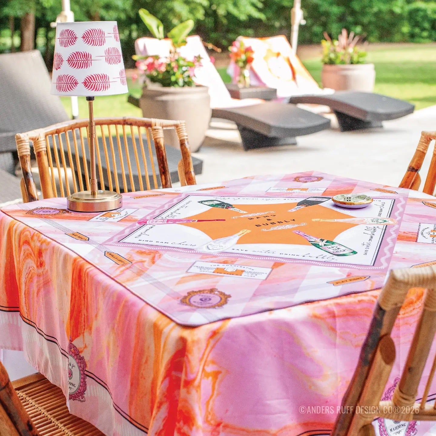 A pink and orange patterned tablecloth with a matching rectangular placemat on a wooden dining table, set in an outdoor patio environment with lounge chairs and plants in the background.
