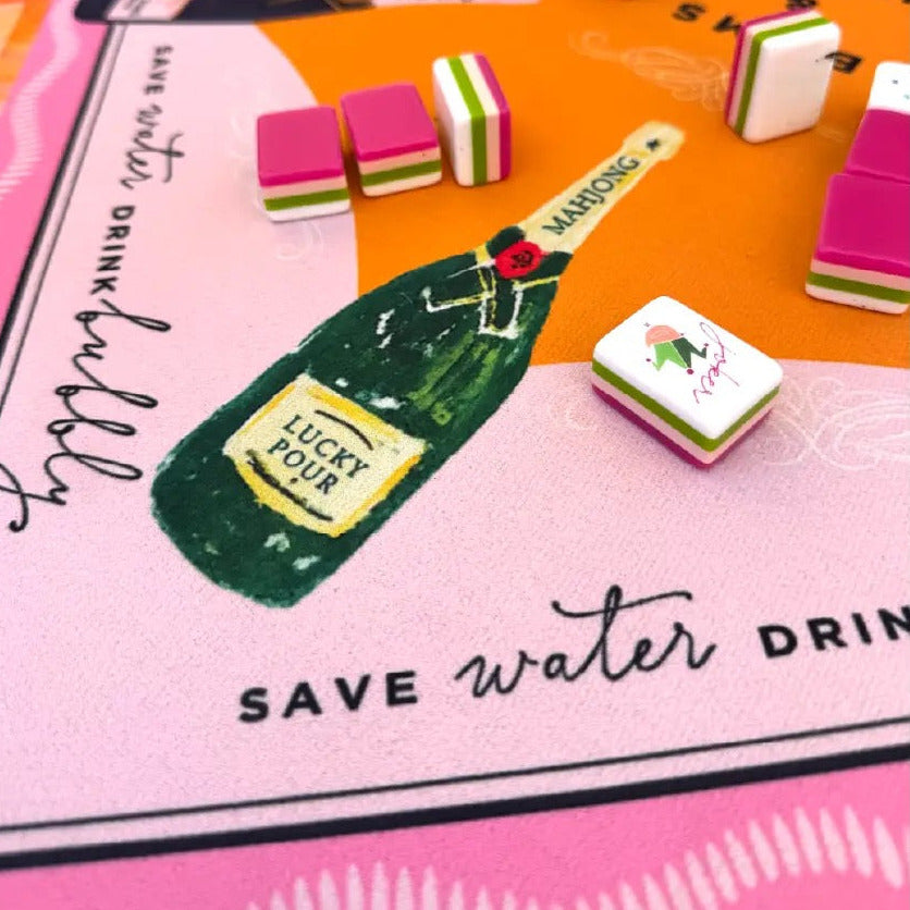 A close-up of a colorful game board featuring a pink and orange design, with small rectangular game pieces and a green bottle illustration. The board includes printed text such as 'SAVE water DRINK bubbly' and 'LUCKY FLOWER'.