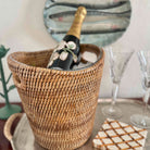 Wicker wine cooler with a bottle of champagne on a table with glasses and a mirror in the background.