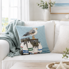 A square decorative throw pillow with a needlepoint-style design of a seagull on a stack of striped beach bags, styled on a white sofa in a bright, modern living room.