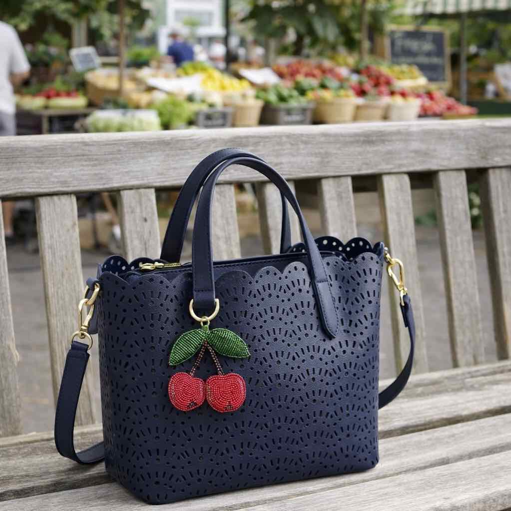 Navy blue handbag with cherry embellishments on a wooden bench at an outdoor market.