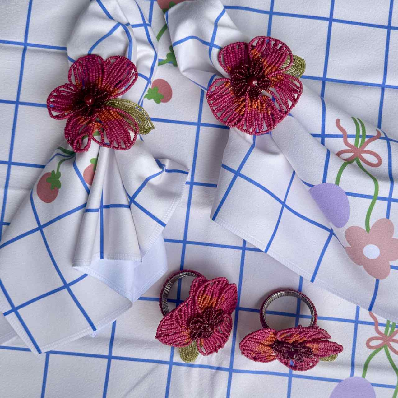 Three pink and red embroidered flower hair clips with green leaves are arranged on a white fabric napkin with a blue grid pattern and a floral print.