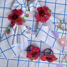 Three pink and red embroidered flower hair clips with green leaves are arranged on a white fabric napkin with a blue grid pattern and a floral print.