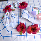 Three beaded flower-shaped napkin rings in pink, red, and orange with green leaves, arranged on a white tablecloth with a blue grid pattern and floral prints.