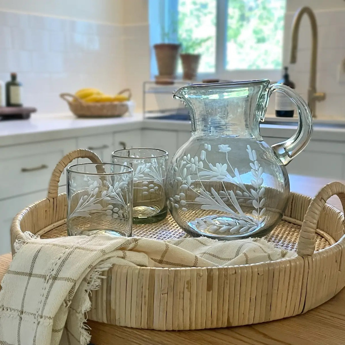 A clear glass pitcher and two matching etched drinking glasses displayed on a woven round tray with a folded cream plaid cloth, set on a wooden table in a bright kitchen.