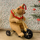 Stuffed toy figurine of a teddy bear wearing a crown and riding a gold-colored bike, with a festive pattern in the background.