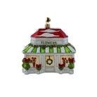 A decorative Christmas ornament shaped like a small flower shop. The white building has a red roof, green-and-white striped awning, red flowers in windows, green holly garland, and a gold hanging loop on top.