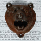 A rustic cast iron bear head bottle opener with a weathered, rusted brown finish, displayed against a background of printed text.