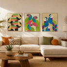 Three framed abstract bird wall art prints hang above a beige sectional sofa in a styled living room. The artwork features colorful toucans, parrots, and peacocks in green, blue, pink, yellow, and black on cream backgrounds.
