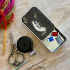 A styled flat-lay of a vintage bird-themed metal sewing box shown open, surrounded by sewing tools including scissors, a spool of black thread, knitting needles, and thread spools on a light wood background.