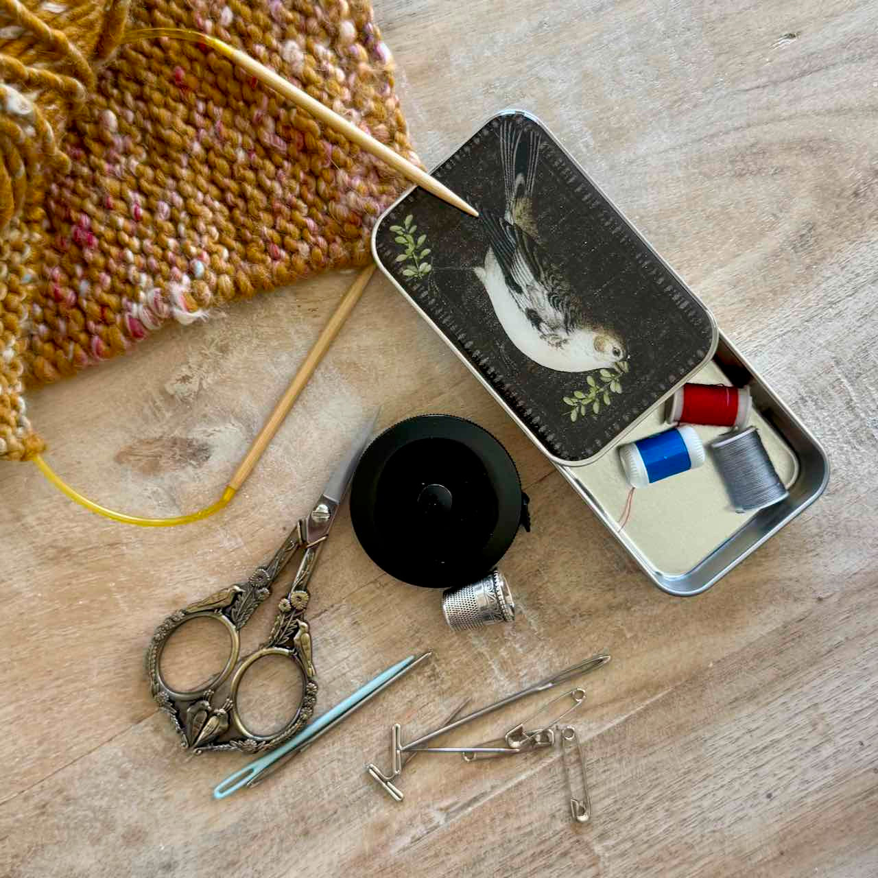 A styled flat-lay of sewing and knitting tools on a light wood surface, including a partially knitted mustard-yellow textile with wooden needles, a small open metal tin with bird artwork and thread spools, a black round container, ornate silver scissors, metal pins, and a thimble.
