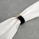 A black ring-shaped napkin holder with a light beige textured end, shown holding a white napkin on a gray textured surface.