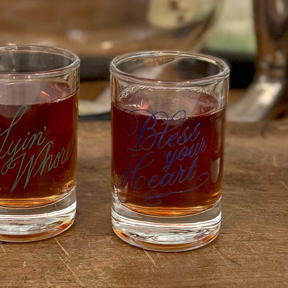 Two clear shot glasses filled with amber liquid on a wooden surface. The right glass has blue script text reading 'Bless your Heart,' while the left glass has teal script text partially visible as 'Yim'Whor.