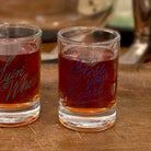 Two clear shot glasses filled with amber liquid on a wooden surface. The right glass has blue script text reading 'Bless your Heart,' while the left glass has teal script text partially visible as 'Yim'Whor.