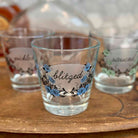 Three clear glass shot glasses with frosted labels and floral designs, displayed on a wooden tray. The center glass is labeled 'blitzed' with blue flowers, and the side glasses are partially visible in the background.