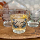 A clear glass cocktail tumbler with blue floral artwork and the word 'blitzed' in script, filled with an amber drink and a lemon wedge, set on a wooden surface with two more matching glasses in the background.