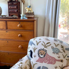 Chair with embroidered design in a room with wooden dresser and window.