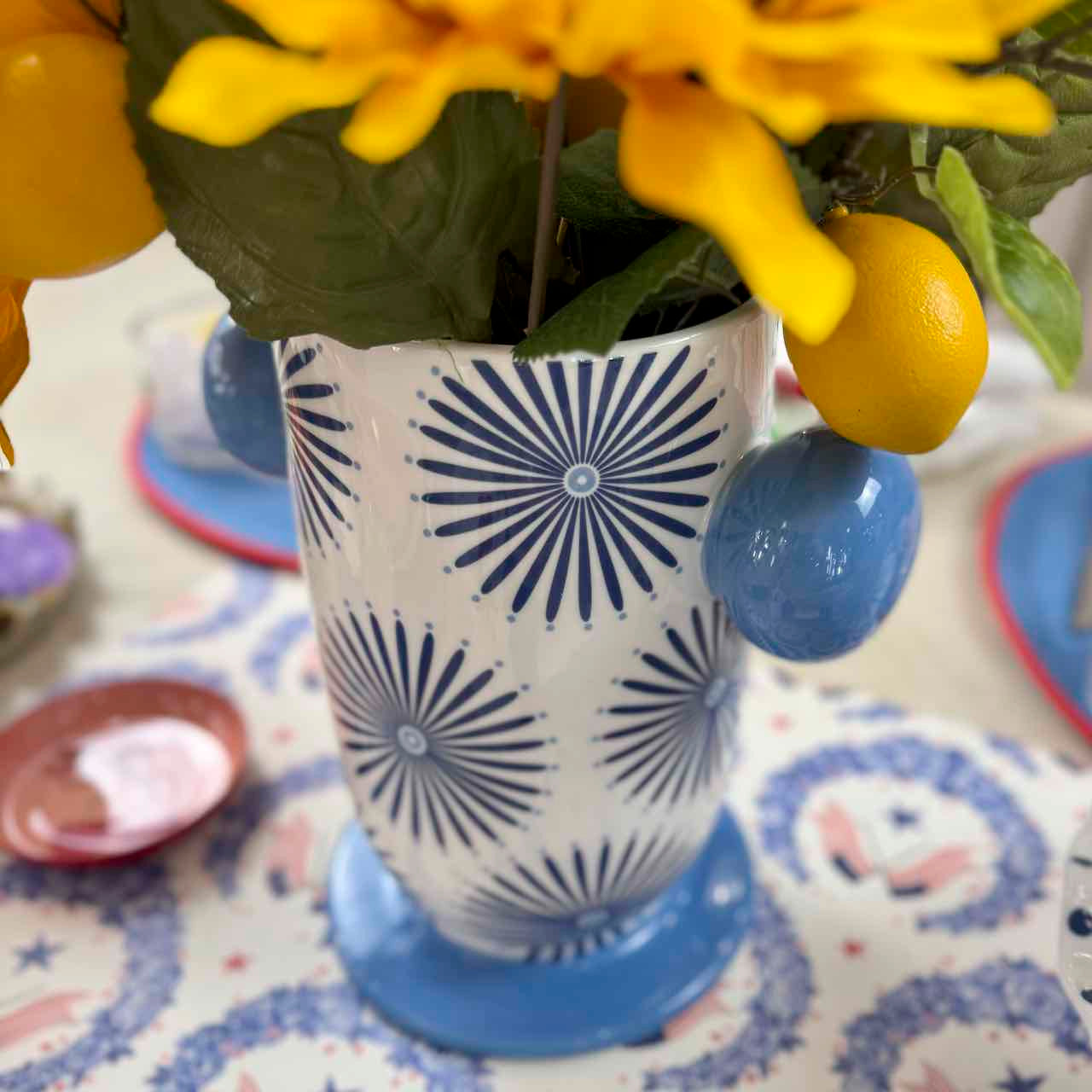 Decorative vase with blue sunburst pattern holding a yellow flower and lemon on a patterned surface.