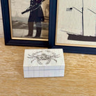 Small wooden box with a crab design on a wooden surface, framed pictures in the background.