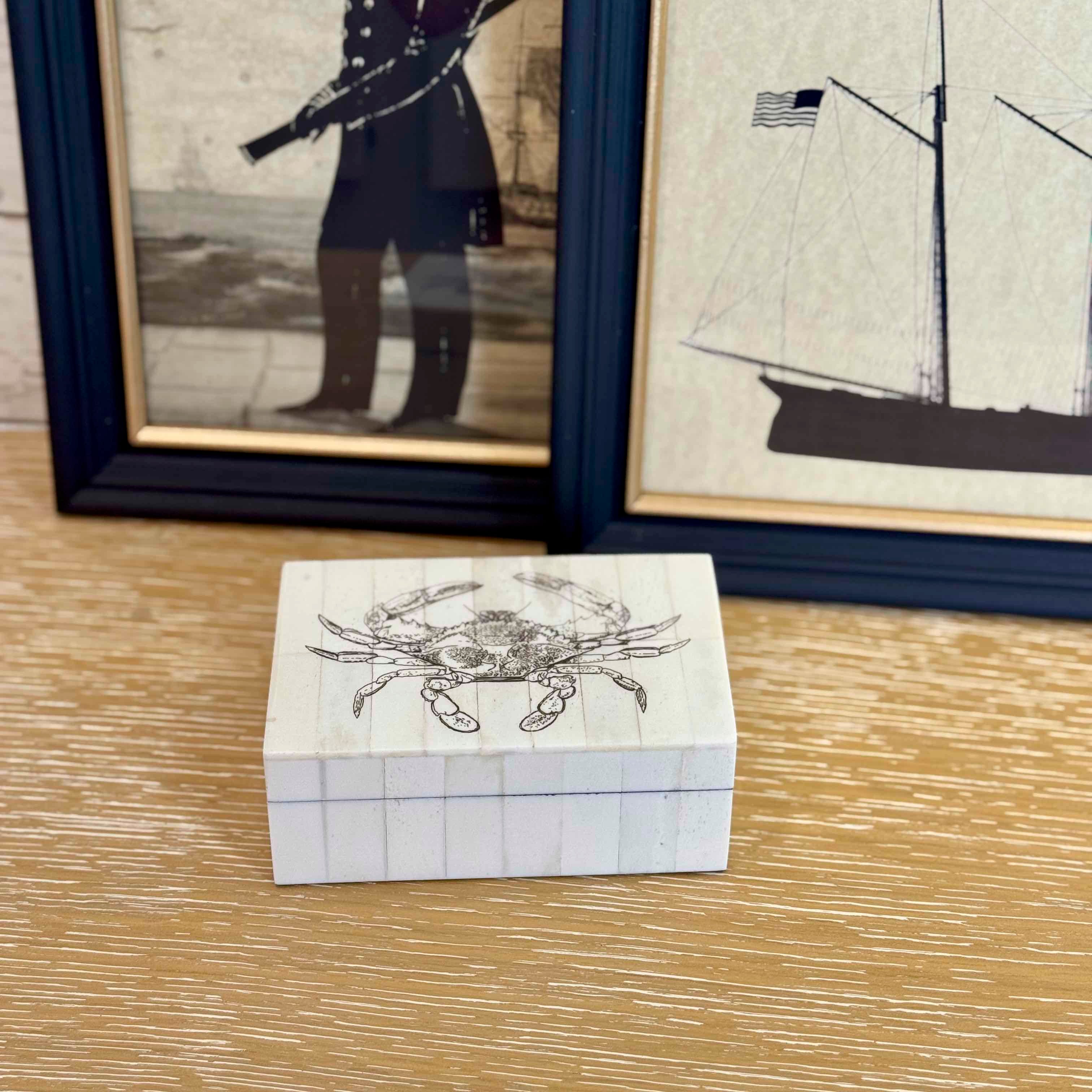Small wooden box with a crab design on a wooden surface, framed pictures in the background.