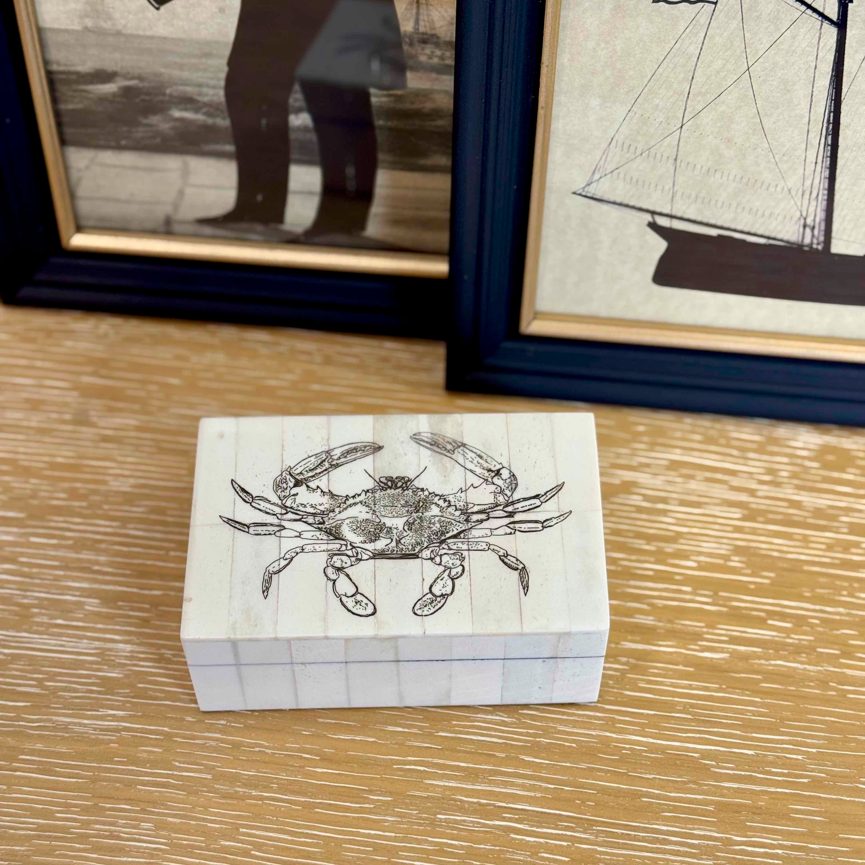 Small wooden box with a crab design on a wooden surface, framed pictures in the background.