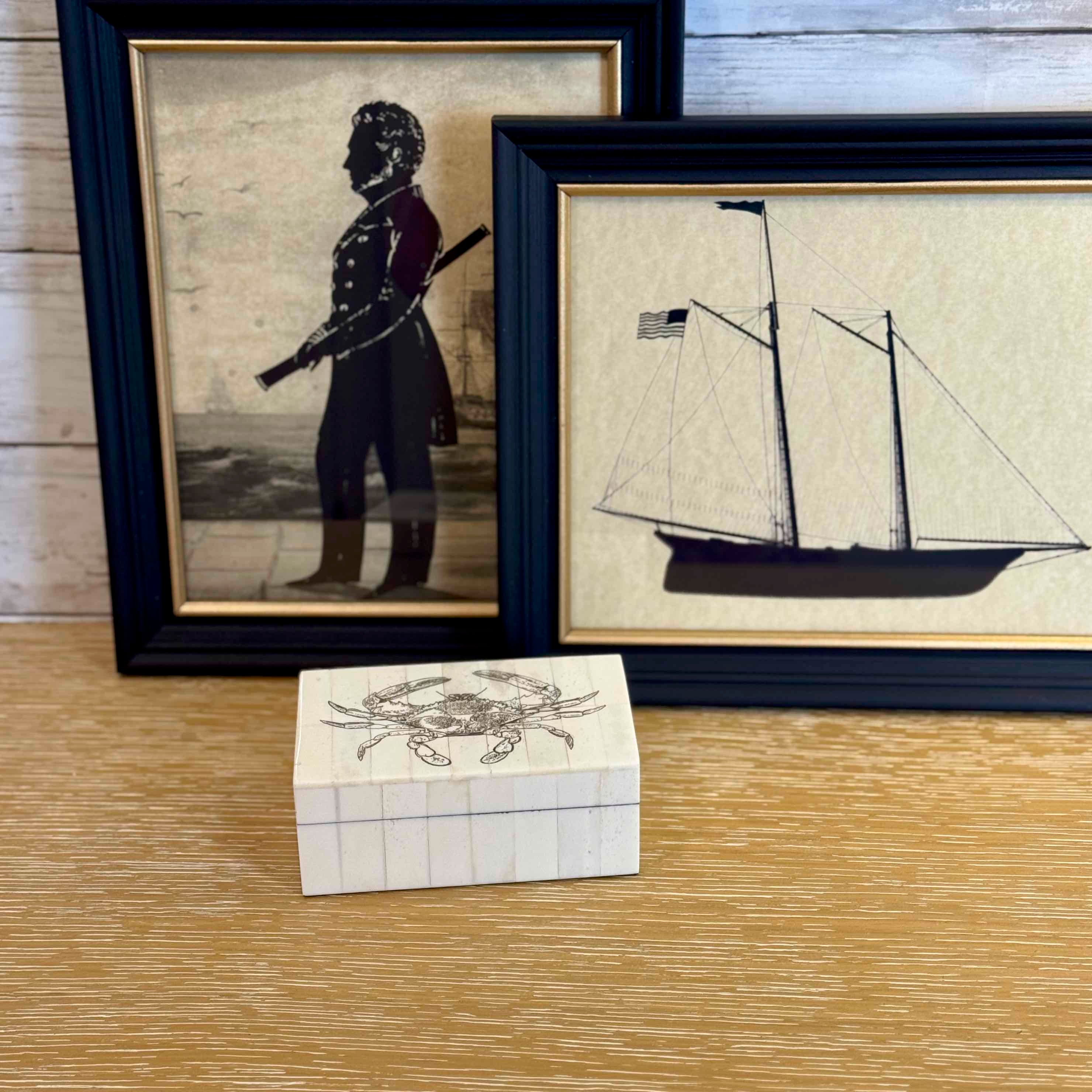 Two framed pictures of a silhouette and a ship, with a small wooden box featuring a crab design, on a wooden surface.