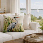 Decorative pillow with a heron design on a white couch in a room with large windows overlooking water.