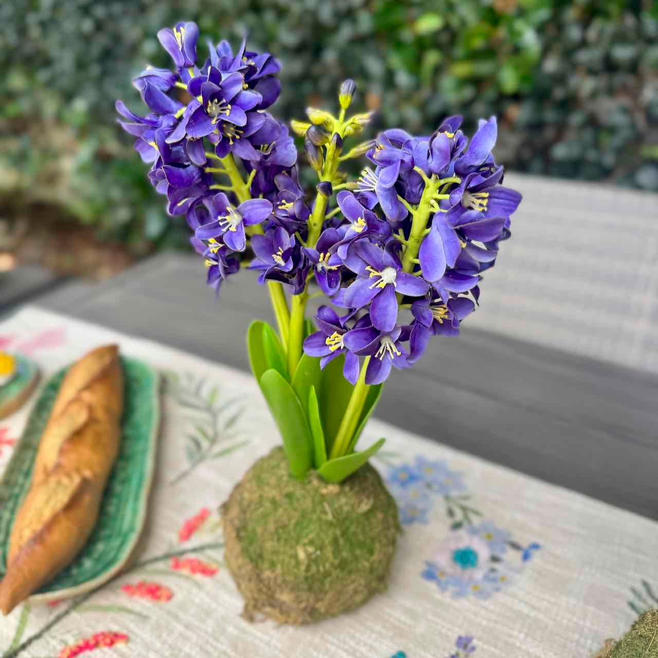 A blooming blue hyacinth flower arrangement in a mossy root vase, set on a floral table runner with a loaf of bread and a green plate in the background.