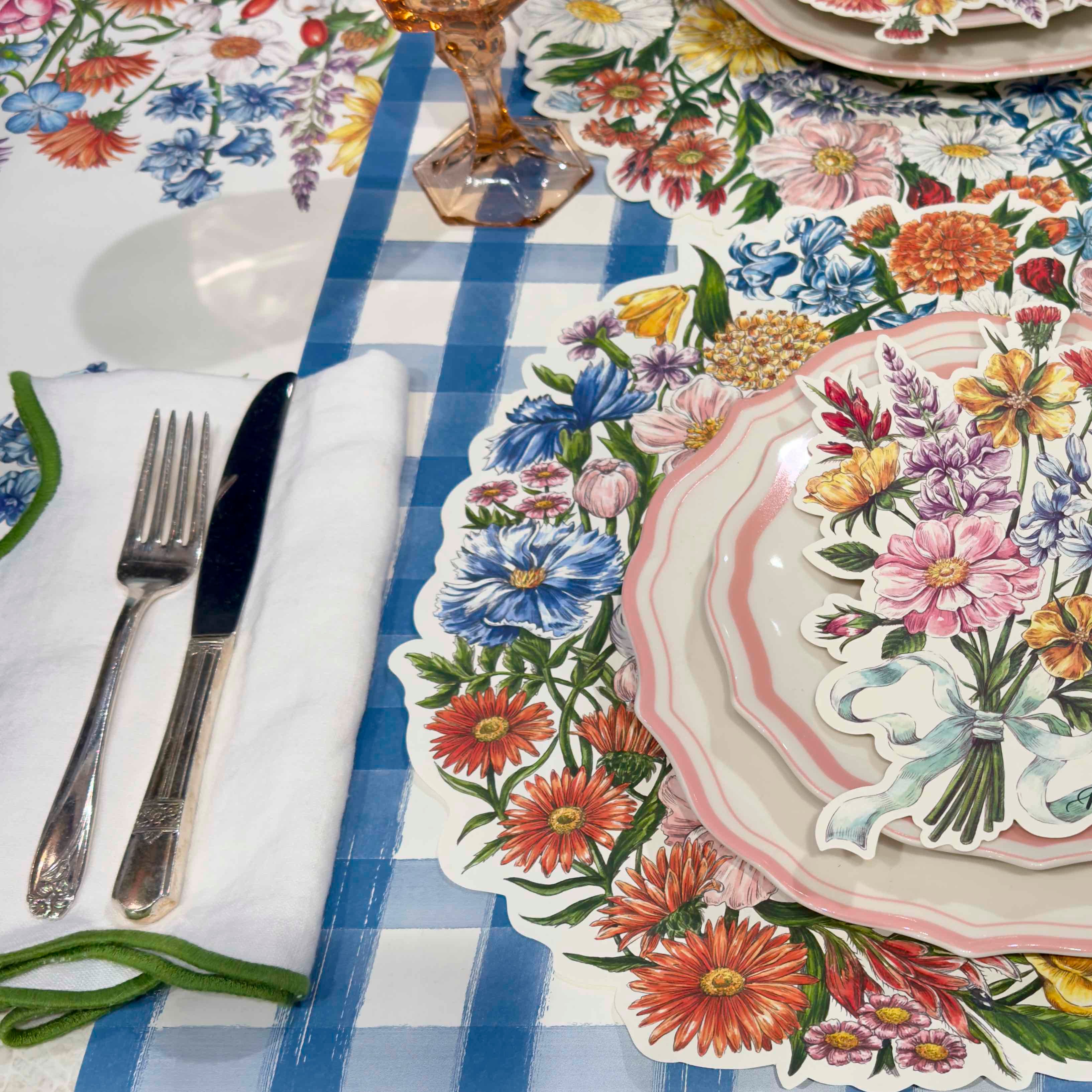 Table setting with floral tablecloth, plates, cutlery, and napkin on a checkered tablecloth.