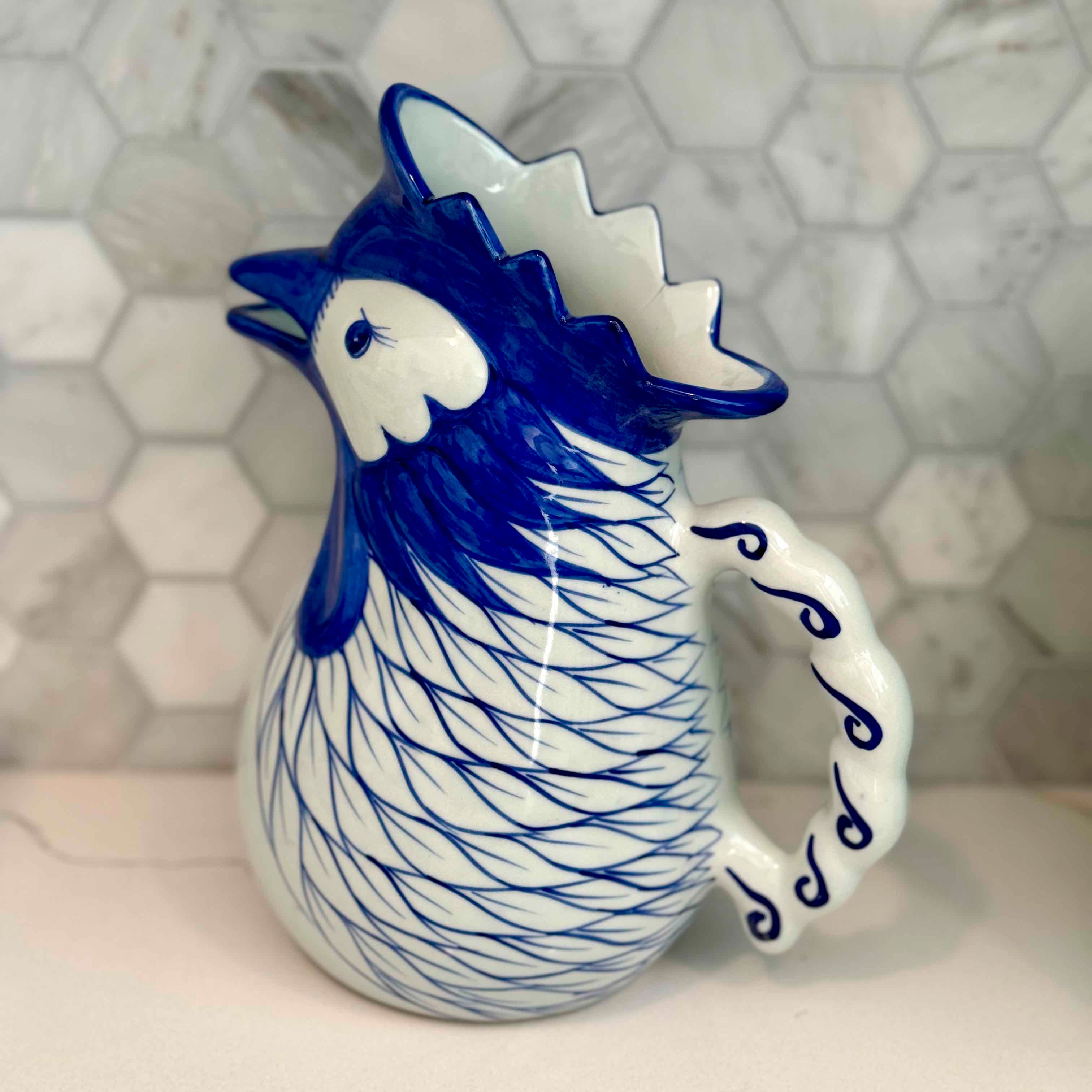 Blue and white ceramic rooster mug on a light gray surface with a geometric patterned wall in the background.