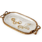 An oval decorative jewelry tray with a ribbed white center and an ornate gold-toned border, shown holding several gold rings and a necklace on a clean white background.