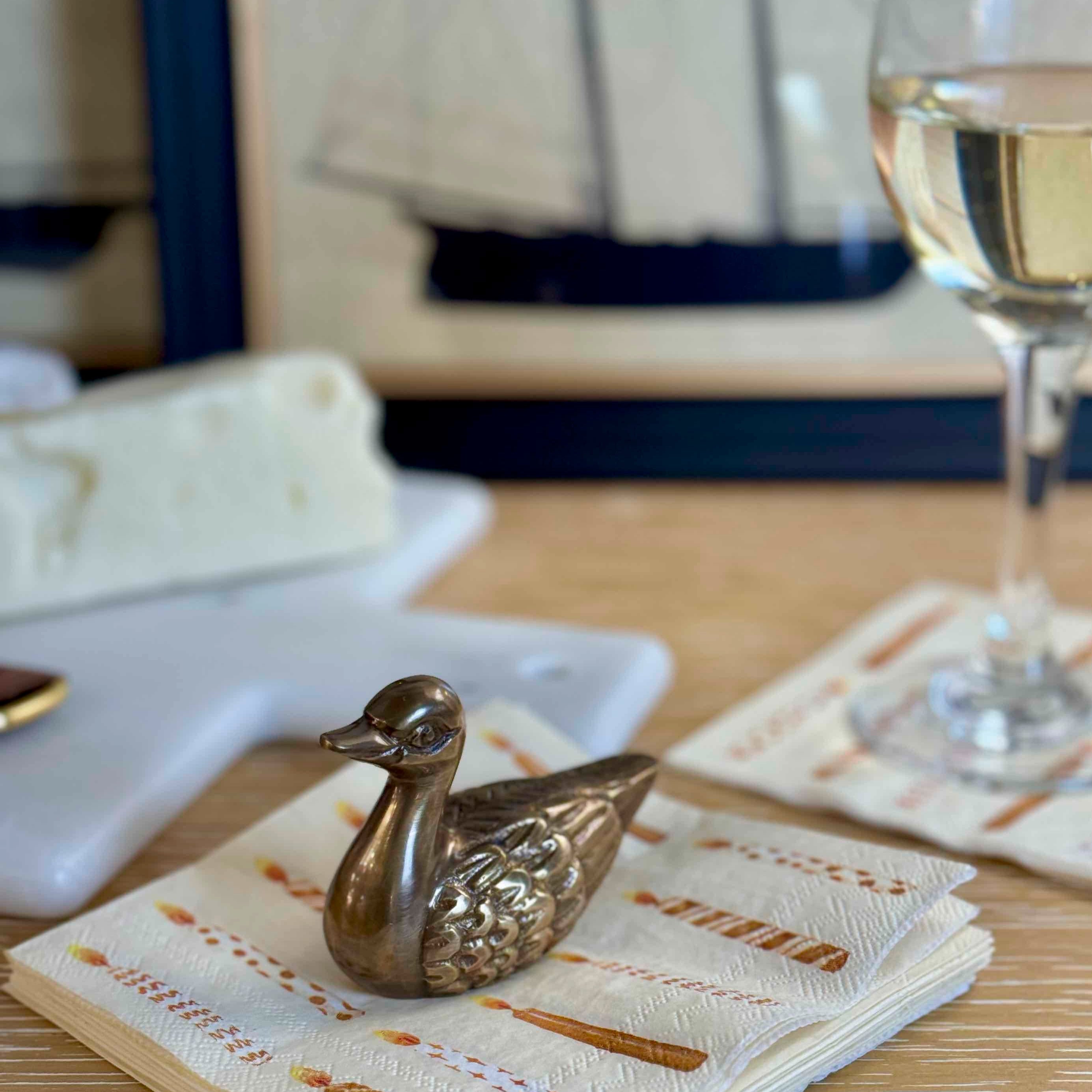 Decorative gold duck on a stack of patterned napkins with a glass of white wine in the background.