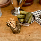 A brass rooster-shaped toothpick holder filled with wooden toothpicks sits on a wooden tabletop next to a small bowl of pickles.