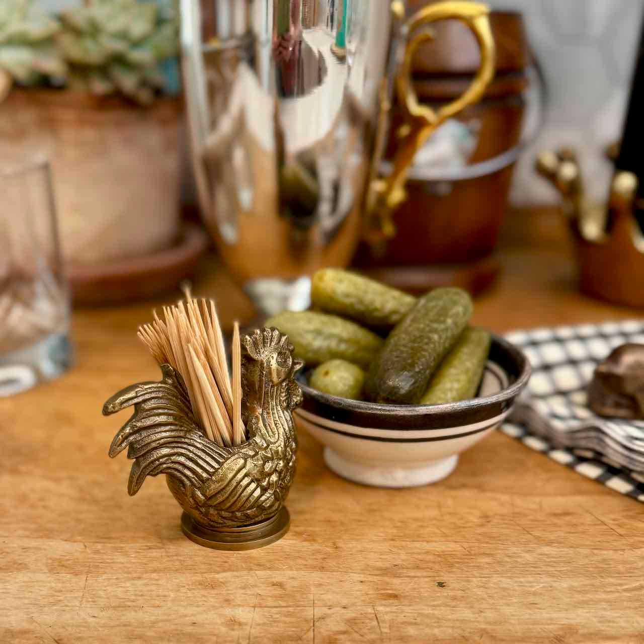 A brass rooster-shaped toothpick holder filled with wooden toothpicks sits on a wooden table next to a small bowl of pickles, with a blurred cocktail shaker and bar decor in the background.