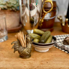 A brass rooster-shaped toothpick holder filled with wooden toothpicks sits on a wooden table next to a small bowl of pickles, with a blurred cocktail shaker and bar decor in the background.