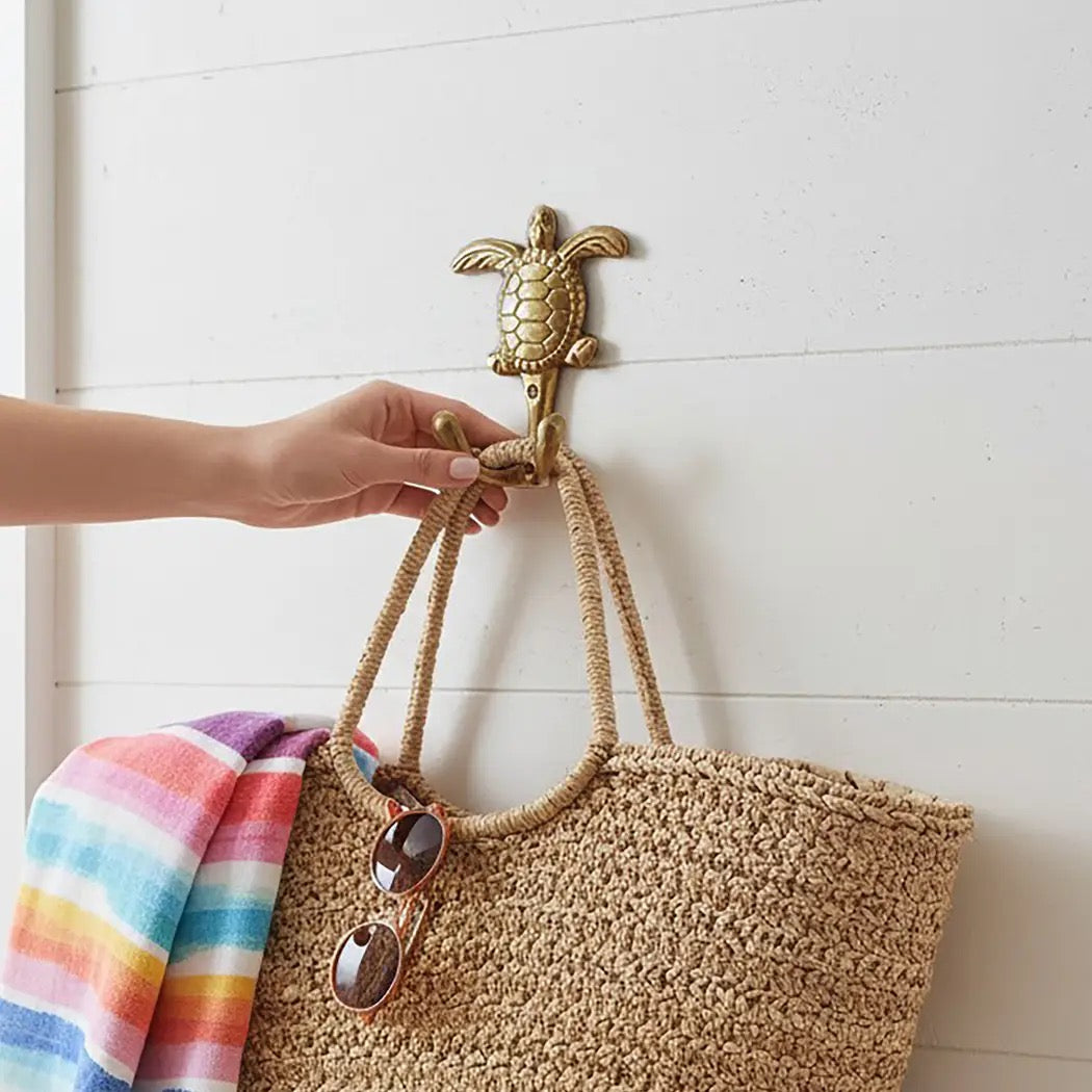 A hand hangs a woven straw tote bag from a gold sea turtle wall hook on a white paneled wall. A striped towel and sunglasses are draped over the bag.