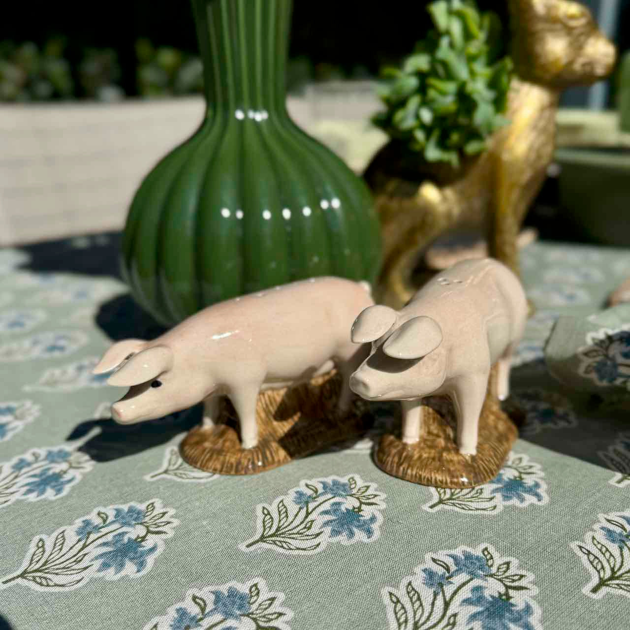 Two ceramic pig figurines on a patterned tablecloth with a green vase and decorative object in the background.