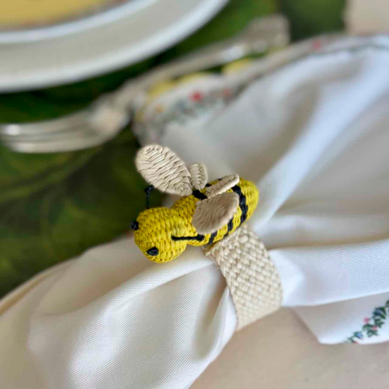 Crocheted bee-shaped napkin ring on a white napkin with a blurred background of cutlery and plates.
