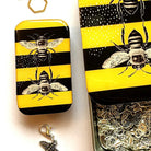 A jewelry box with a yellow and black striped design featuring black and white illustrations of bumblebees. The box is shown open, revealing silver-toned metal chain links inside, with a small butterfly charm dangling below.