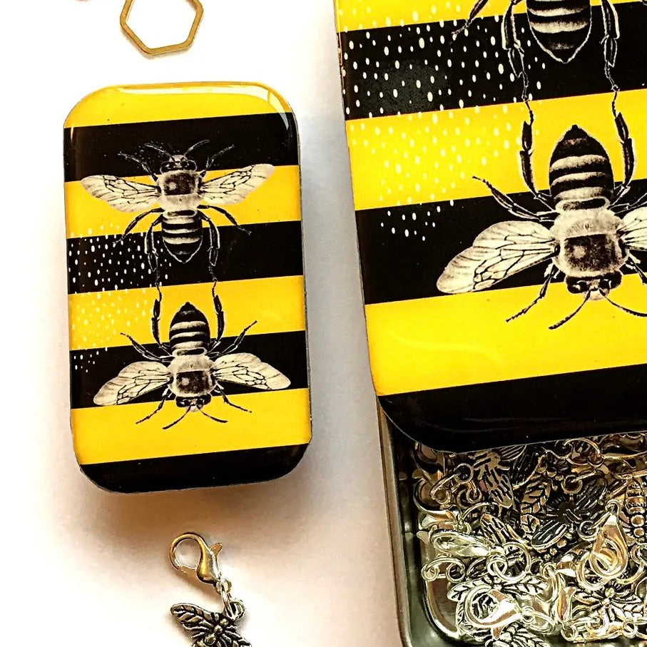A jewelry box with a yellow and black striped design featuring black and white illustrations of bumblebees. The box is shown open, revealing silver-toned metal chain links inside, with a small butterfly charm dangling below.