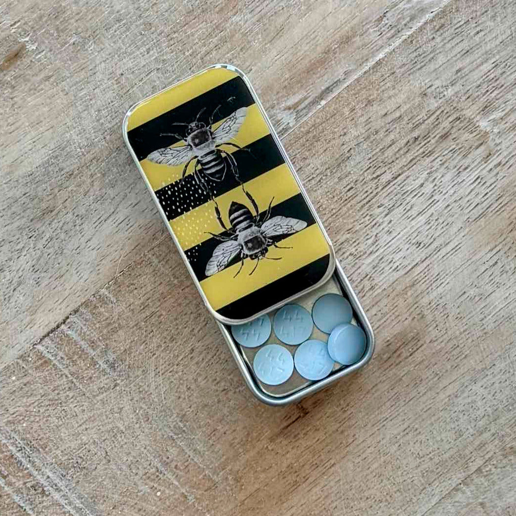 An open silver metal pill box with a yellow and black striped lid decorated with two bees, containing light blue round tablets, placed on a light wood surface.
