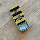 An open silver metal pill box with a yellow and black striped lid decorated with two bees, containing light blue round tablets, placed on a light wood surface.