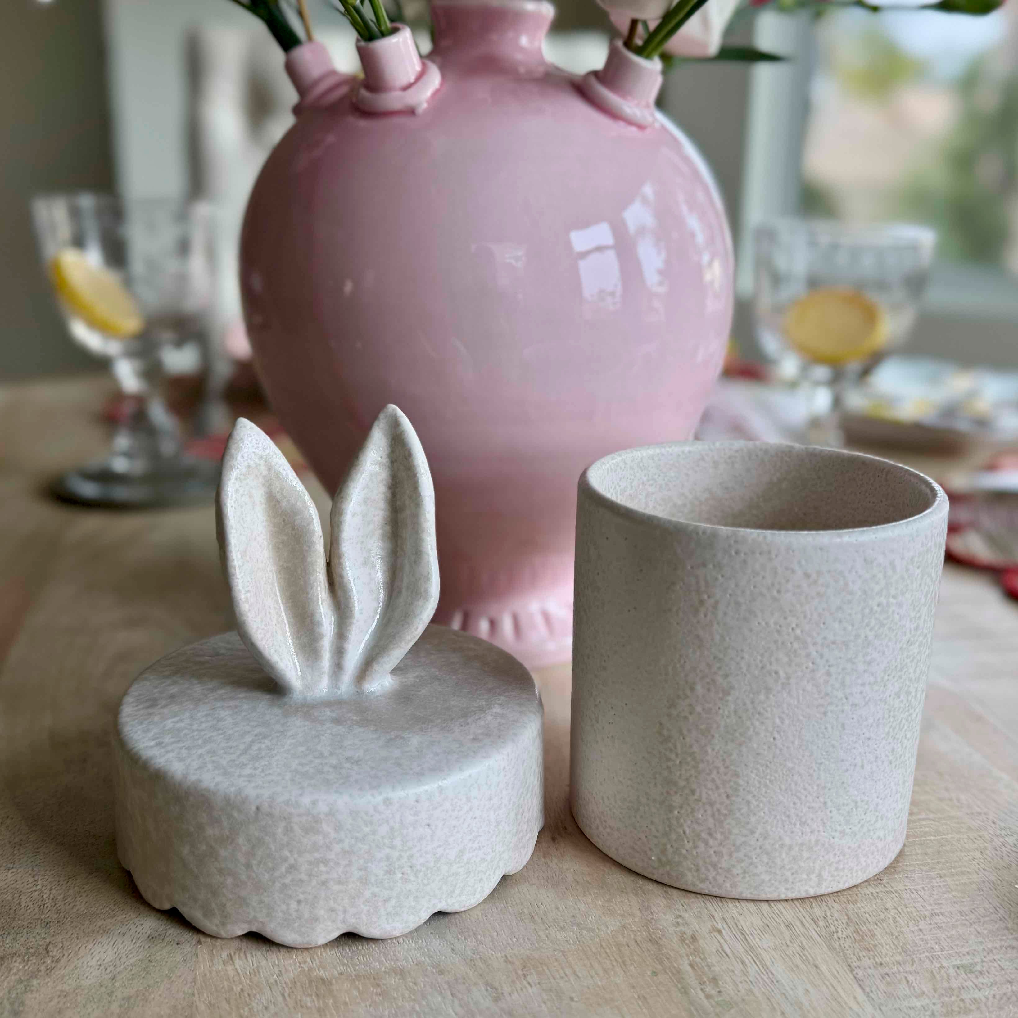 Decorative items including a pink vase, a small white container with rabbit ears, and a textured white cup on a table.